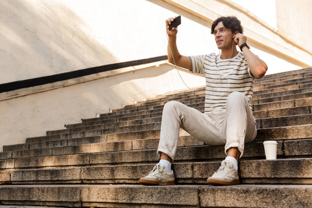 Image of handsome young man sitting on steps outdoors listening music take a selfie by mobile phone. - Foto, immagini