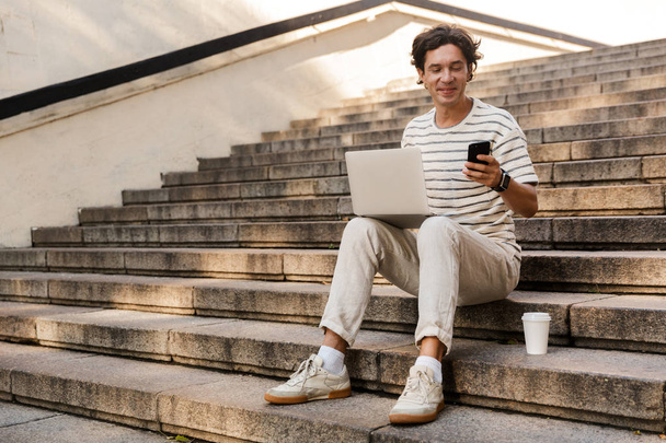 Image of handsome young man sitting on steps outdoors using laptop computer and mobile phone. - Foto, Imagen