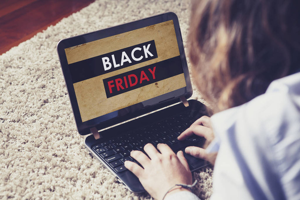 Black Friday on line store in a laptop screen while woman uses it to buy, lying down on the carpet at home. - Photo, image