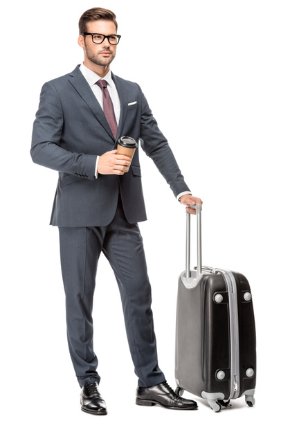 joven hombre de negocios guapo con el equipaje y la taza de papel de café mirando hacia otro lado aislado en blanco
 - Foto, imagen