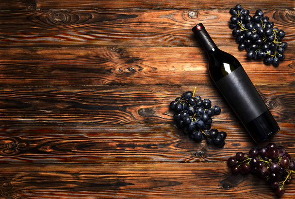 Unopened vintage bottle of red wine with blank matte black label & bunches of ripe grapes on wooden table background. Expensive bottle of cabernet sauvignon concept. Copy space, top view flat lay - Фото, изображение