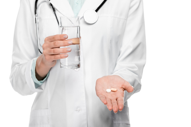 abgeschnittenes Bild einer Ärztin in medizinischem Mantel, die ein Glas Wasser und Tabletten auf weiß isoliert hält - Foto, Bild