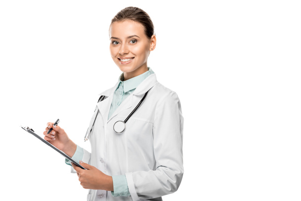 sonriente joven doctora en abrigo médico escrito en portapapeles aislado en blanco
 - Foto, Imagen