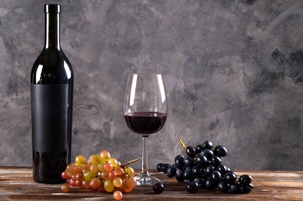 Vintage bottle of red wine with blank matte black label, bunch of grapes on wooden table, concrete wall background. Expensive bottle of cabernet sauvignon concept. Copy space, top view, flat lay. - Foto, imagen