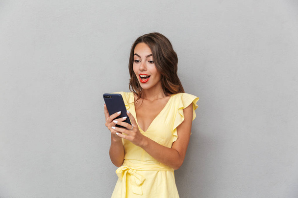 Portrait of a joyful young girl in dress standing over gray background, holding mobile phone - Foto, Imagen