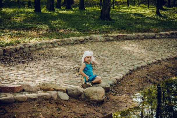 Little girl in white hat and blue dress sitting on stone bank of river in park - Zdjęcie, obraz