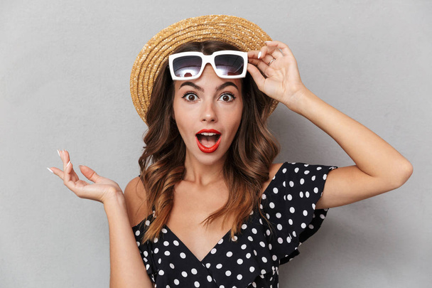 Portrait of an excited young girl in dress and straw hat over gray background, posing with sunglasses - Photo, Image