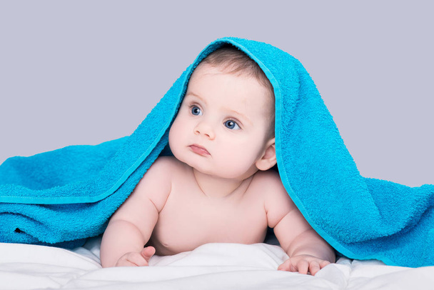 Serious baby girl or boy with big blue eyes lying on belly under the blue towel after shower, isolated over gray background - Foto, Imagen