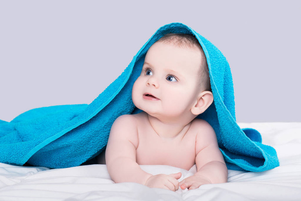 Surprised smiling baby girl or boy with big blue eyes lying on belly under the blue towel after shower, isolated over gray background - Foto, immagini