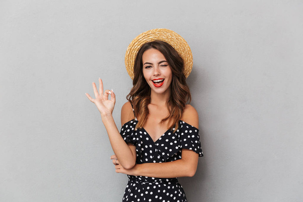 Portrait of a cheerful young girl in dress and straw hat over gray background, showing ok gesture - Foto, Imagen