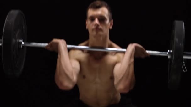 Man is doing exercises with a barbell, is engaged in a crossfit on a black background in the studio - Video