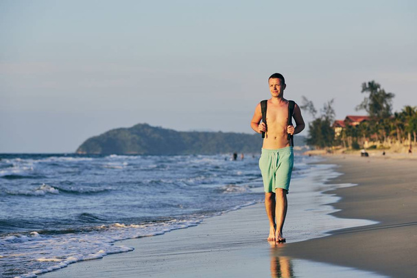 Hombre joven (turista) con mochila caminando en la playa de arena
. - Foto, imagen