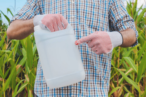 Farmer holding pesticide chemical jug in cornfield. Blank unlabelled bottle as mock up copy space for herbicide, fungicide or insecticide used in corn crop farming. - 写真・画像