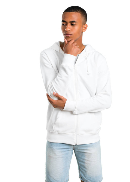 Dark-skinned young man with white sweatshirt standing and looking to the side with the hand on the chin on isolated white background - Photo, Image