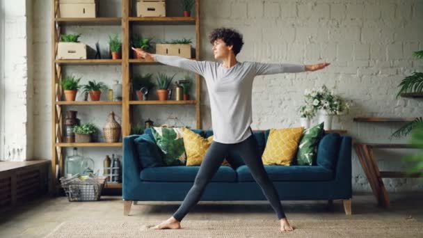 Beautiful girl is doing yoga exercises at home stretching body standing at home on floor concentrated on practice. Young people, interiors and healthy lifestyle concept. - 映像、動画