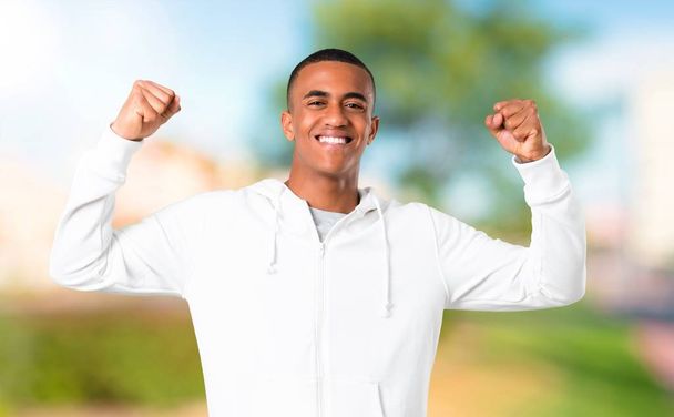 Dark-skinned young man with white sweatshirt celebrating a victory in winner position in a park - Foto, Imagem