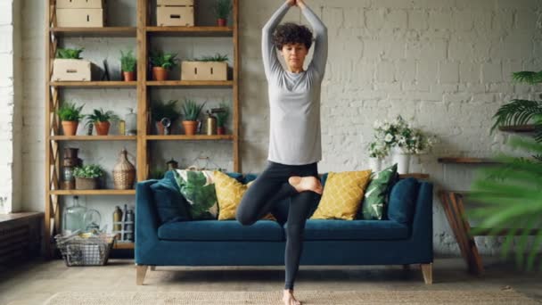 Pretty girl is focused on yoga practice at home doing balancing exercises standing on floor on one leg. Healthy lifestyle, youth and apartment concept. - Filmati, video