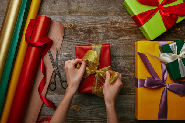 cropped shot of woman tying ribbon on wrapped christmas present on wooden surface with other gifts  - Foto, Bild