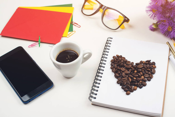 Schreibtisch mit Tasse Kaffee. Arbeitsbereich mit Notebook, Bürobedarf, Brille, Smartphone und Kaffeetasse. Kaffeebohnen auf Papier. moderner weißer Bürotisch. Kaffeepause am Arbeitstisch - Foto, Bild