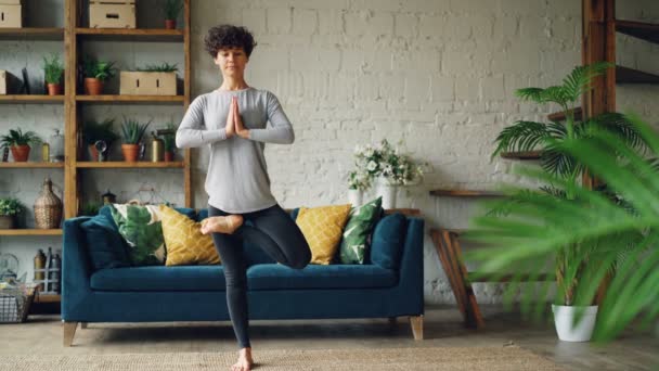 Attractive young woman is balancing on one leg in Tree position with hands in Namaste during yoga at home alone. Healthy people, flats and sports concept. - Felvétel, videó