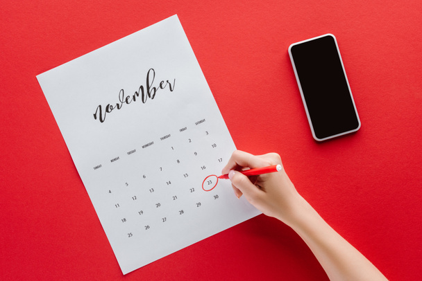 обрезанный вид женщины, пишущей черную пятницу в календаре на ноябрь изолированы на красный с помощью смартфона
 - Фото, изображение