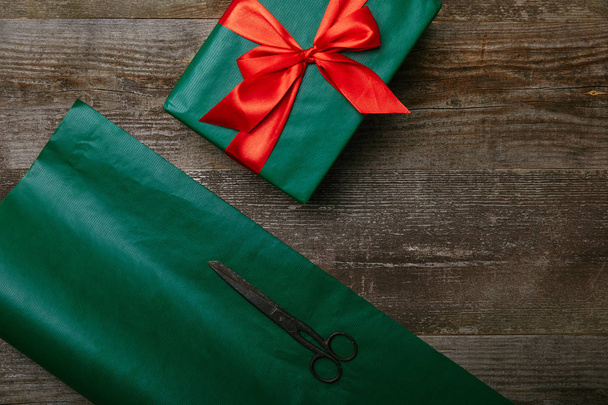 top view of green wrapping paper, scissors and present with red ribbon on wooden background - Fotoğraf, Görsel