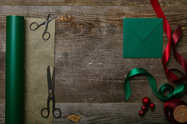 flat lay with blank envelope, scissors, green and red ribbons and wrapping paper on wooden surface, christmas background - Photo, Image