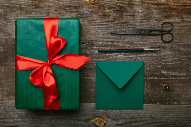 lapos feküdt becsomagolt karácsonyi ajándék piros szalag, olló és boríték üdvözlőlap fa felületre - Fotó, kép