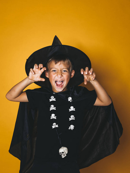 boy dressed in halloween witcher costume with black cap on head showing frightening gesture and looking at camera on orange background - Φωτογραφία, εικόνα