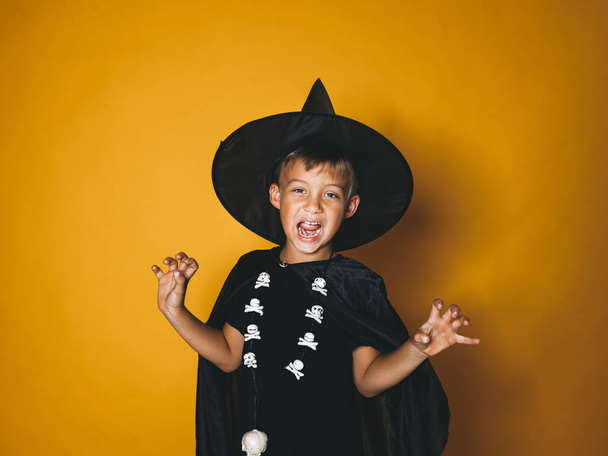 boy dressed in halloween witcher costume with black cap on head showing frightening gesture and looking at camera on orange background - Photo, Image