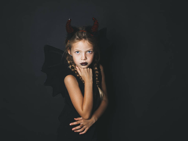 beautiful blond girl dressed in halloween devil costume with red horns and wings looking at camera on dark background - Photo, image