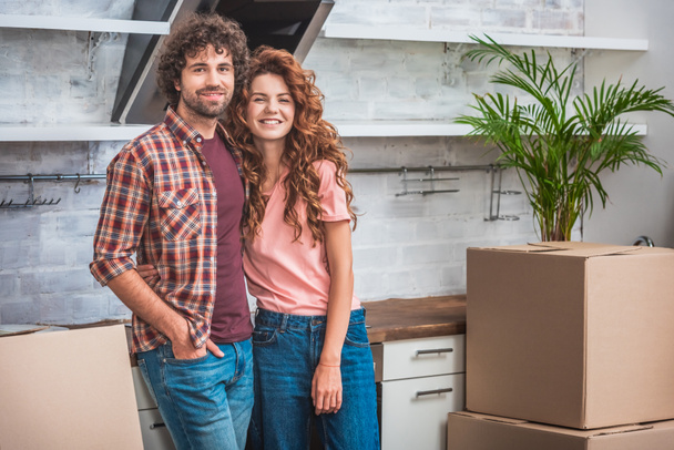 smiling couple with curly hair standing near cardboard boxes and looking at camera at new kitchen - Фото, изображение