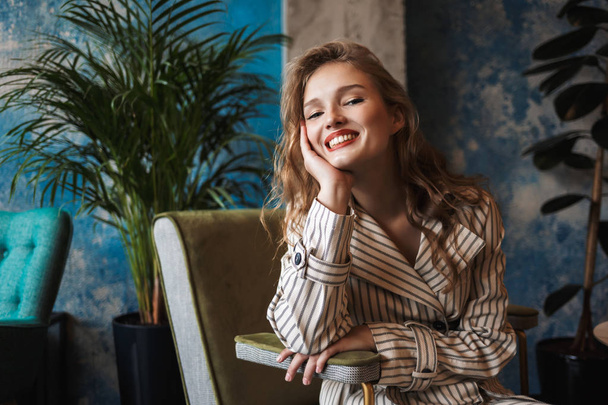 Young cheerful woman with wavy hair in striped trench coat leaning on hand happily looking in camera spending time in modern cafe - Фото, изображение