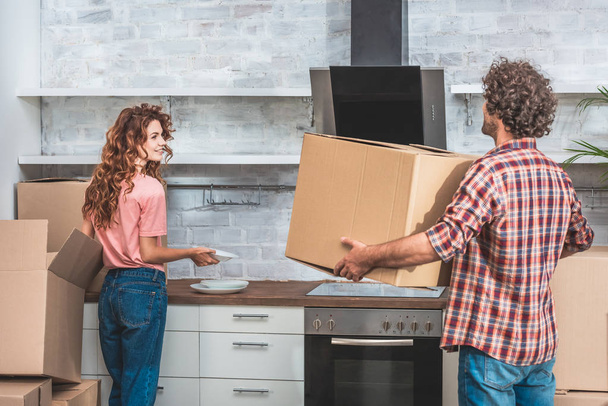 boyfriend and girlfriend with curly hair unpacking cardboard boxes together at new kitchen - Foto, afbeelding