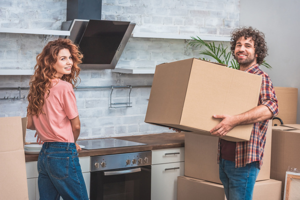 smiling couple unpacking cardboard boxes together at new kitchen and looking at camera - Photo, Image