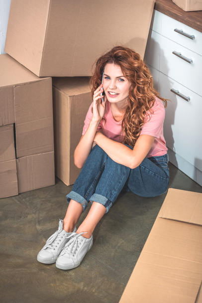 mujer hermosa feliz con el pelo rizado sentado en el suelo cerca de cajas de cartón y hablando por teléfono inteligente en el nuevo hogar
 - Foto, Imagen