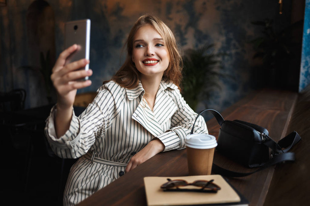 Fiatal vidám nő csíkos viharkabát boldogan fotózni a kis fekete táska és kávé cellphone közel modern kávézó időt menni - Fotó, kép