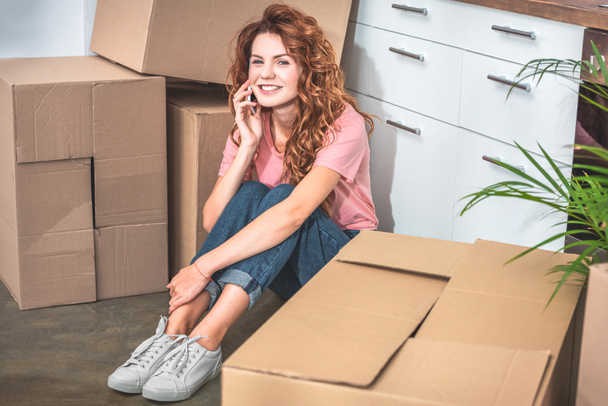 smiling attractive woman sitting on floor near cardboard boxes and talking by smartphone at new home - Фото, зображення
