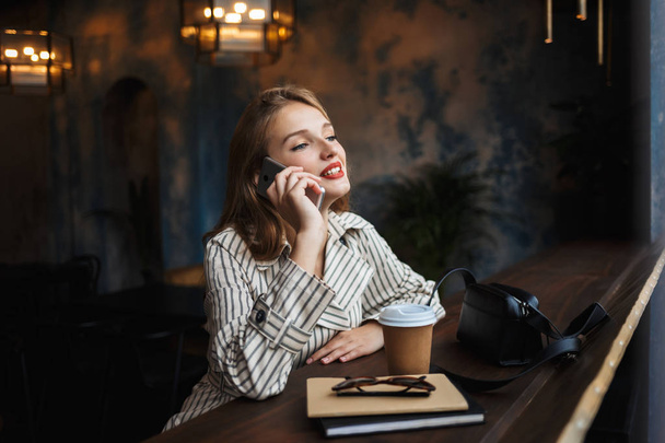 Beautiful smiling girl in striped trench coat talking on cellphone  dreamily looking aside with little black handbag and coffee to go near spending time in modern cafe - Foto, Imagem