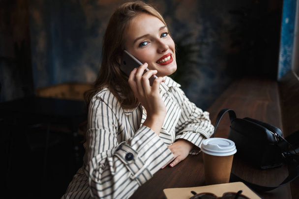 Hermosa dama sonriente en gabardina rayada hablando en el teléfono celular mirando felizmente en la cámara con pequeño bolso negro y café para ir cerca de pasar tiempo en la cafetería moderna
 - Foto, Imagen