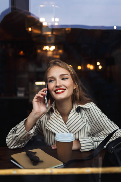 Chica alegre en gabardina rayada hablando en el teléfono celular mirando a un lado con una taza de café para ir cerca de pasar tiempo en la cafetería acogedora moderna
 - Foto, Imagen