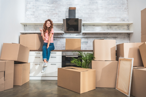 smiling attractive woman sitting on kitchen counter between cardboard boxes and showing peace sign at new home - Photo, Image