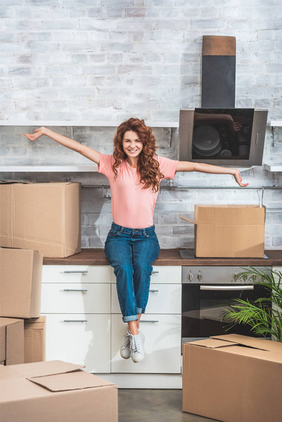happy beautiful woman sitting on kitchen counter with outstretched hands between cardboard boxes at new home - Foto, Imagen