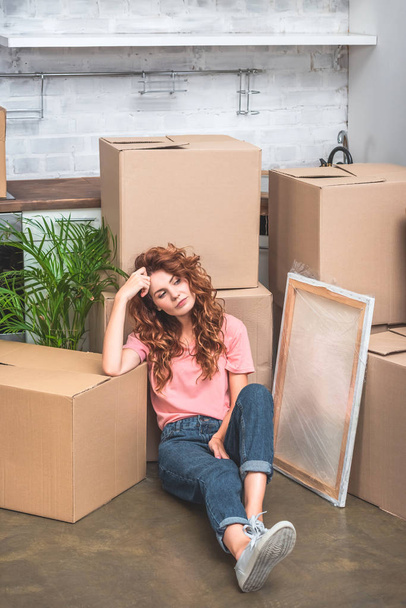 high angle view of attractive woman with curly red hair sitting on floor near cardboard boxes at new home - Photo, Image