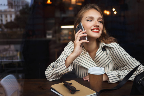 Jeune femme souriante en trench coat rayé parlant sur téléphone portable regardant joyeusement de côté avec une tasse de café pour aller près de passer du temps dans un café confortable moderne
 - Photo, image