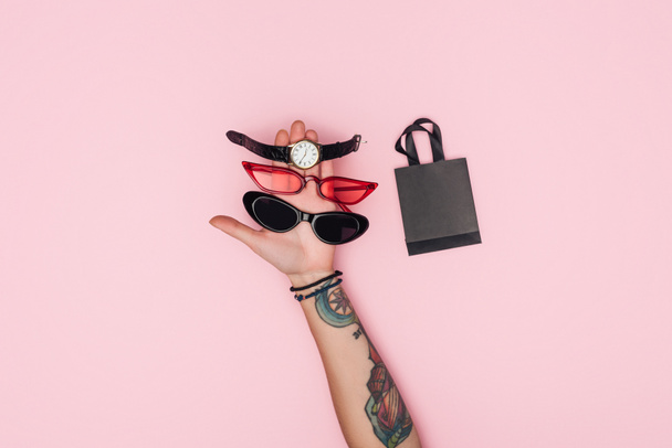 abgeschnittene Ansicht tätowierter Frau mit trendiger Sonnenbrille und Uhr in der Nähe kleiner Einkaufstasche isoliert auf rosa - Foto, Bild