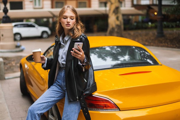 Young attractive woman in leather jacket leaning on yellow sport car holding cup of coffee to go in hand thoughtfully using cellphone on city street - Foto, imagen