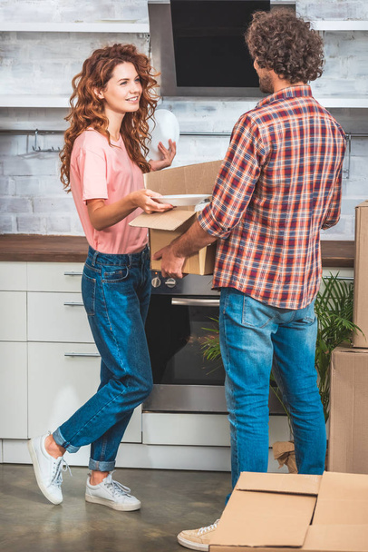 pareja desempacando utensilio de la caja de cartón en la cocina nueva y mirándose entre sí
 - Foto, imagen
