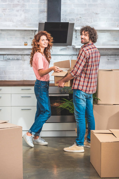smiling couple unpacking cardboard box together at new home and looking at camera - Foto, Bild