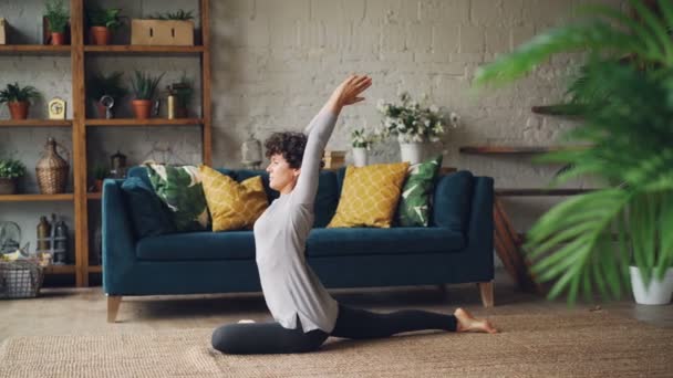 Attractive young woman is stretching legs and arms sitting on floor during individual practice at home. Healthy lifestyle, interior and people concept. - Filmagem, Vídeo